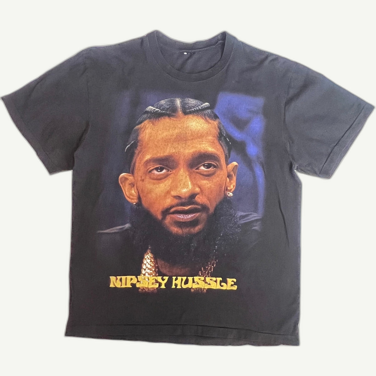 A faded black graphic t-shirt featuring a portrait of rapper Nipsey Hussle with his name printed in yellow text below the portrait, laid flat against a light off-white background.