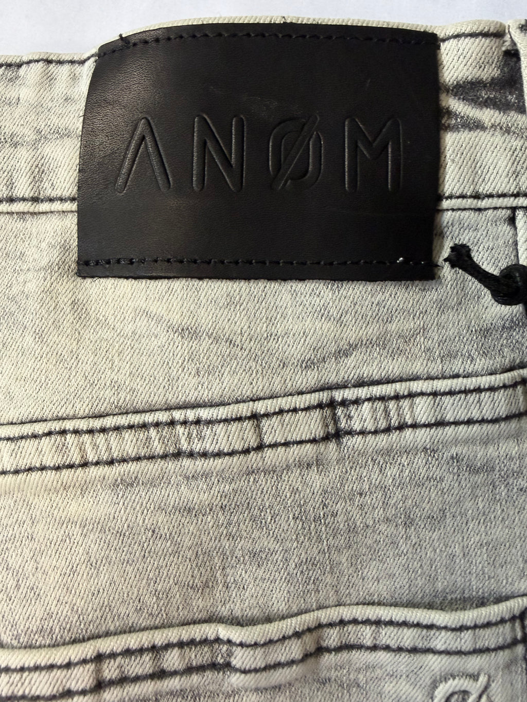 Macro detail view of a premium black leather waistband patch on AMNÉ modern streetwear denim jeans. Features a debossed geometric brand logo over a heavily textured grey acid wash canvas.