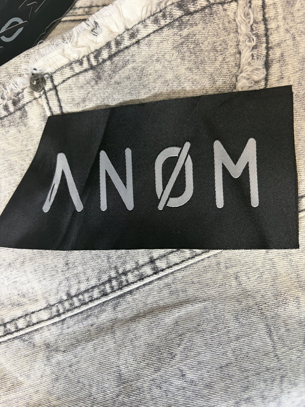 Detailed macro view of branded textile tags on AMNÉ modern streetwear denim jeans. Features a black pocket patch overlay with a custom silver logo over heavily marbled charcoal acid wash fabric.