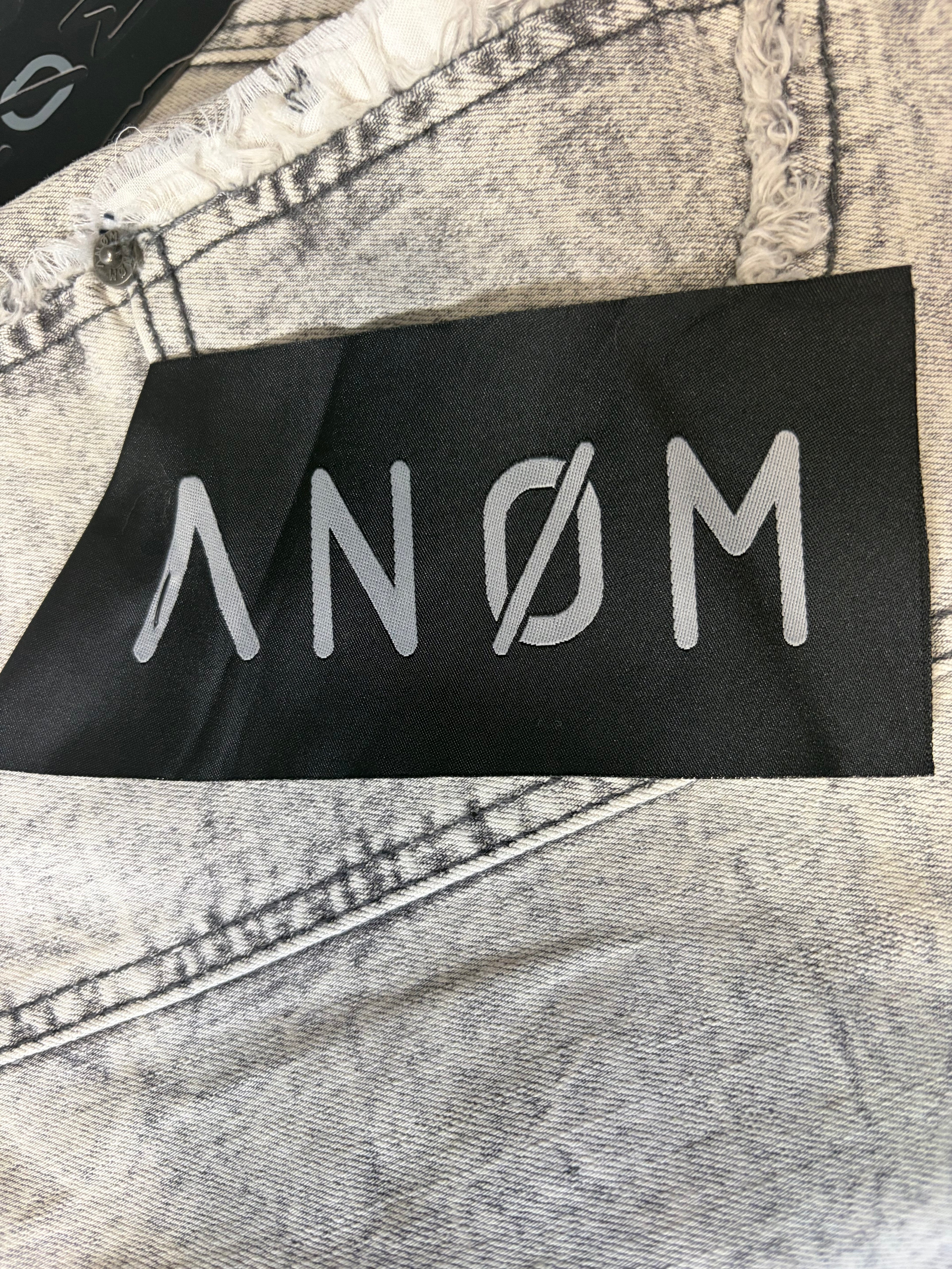 Detailed macro view of branded textile tags on AMNÉ modern streetwear denim jeans. Features a black pocket patch overlay with a custom silver logo over heavily marbled charcoal acid wash fabric.