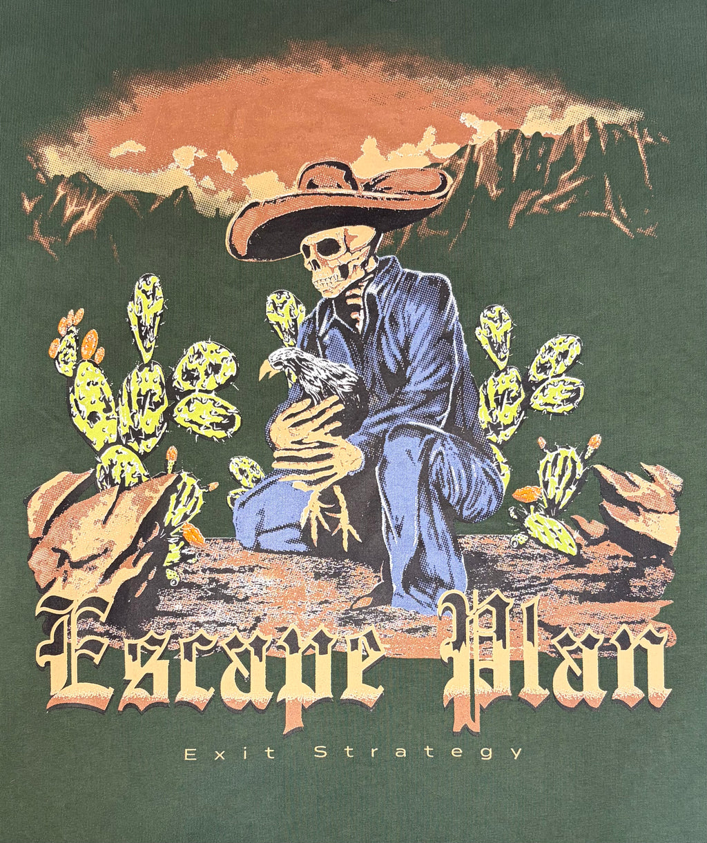 A graphic print on a dark green shirt featuring a skeleton cowboy in a blue suit kneeling among cacti under a mountain sunset sky, with the text 'Escape Plan' and 'Exit Strategy'.