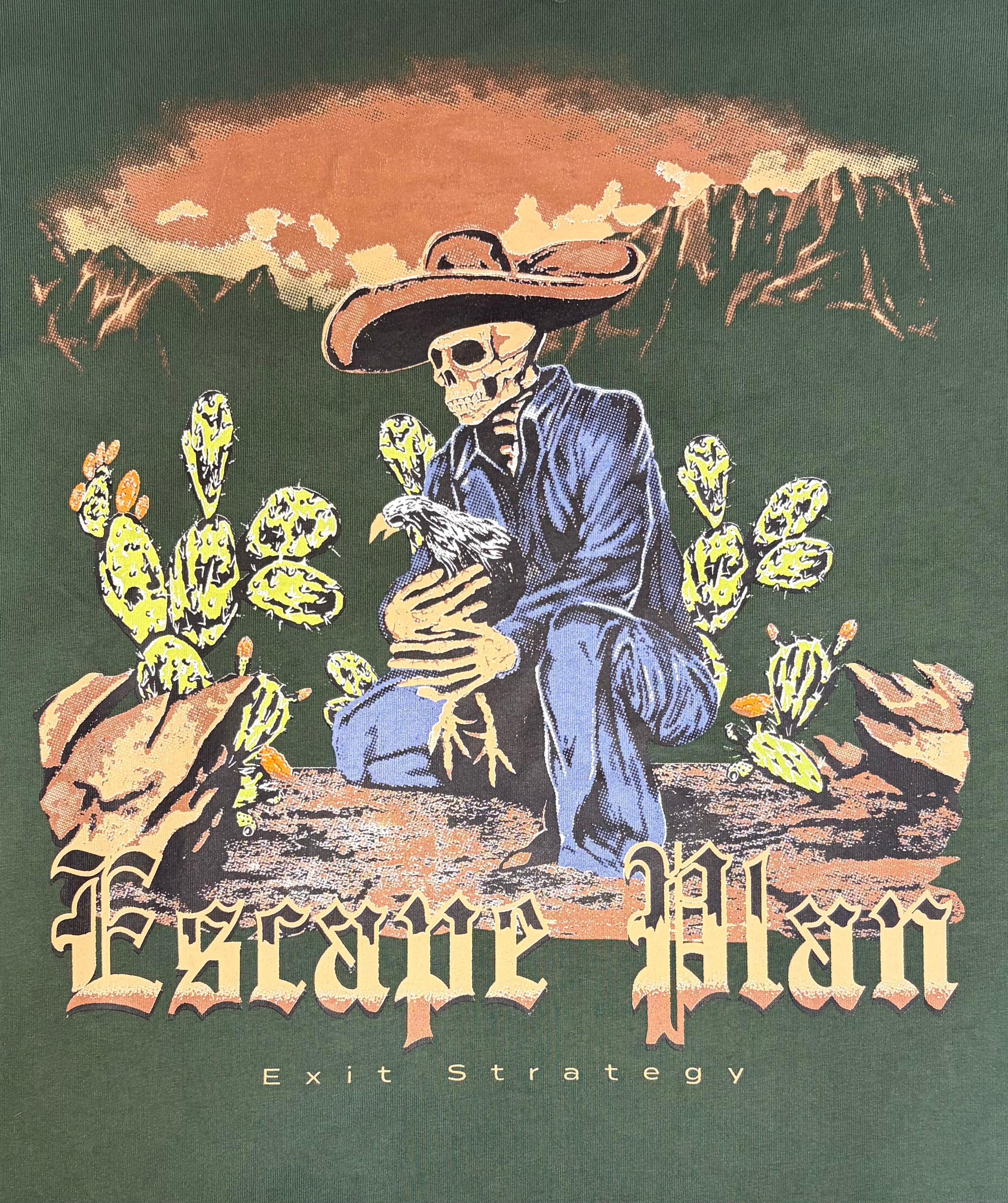 A graphic print on a dark green shirt featuring a skeleton cowboy in a blue suit kneeling among cacti under a mountain sunset sky, with the text 'Escape Plan' and 'Exit Strategy'.