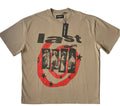 A beige Last Saints short sleeve graphic T-shirt featuring bold black typography that reads "last saint" layered over a four-panel religious portrait artwork. The graphic includes a large, distressed red circular swirl pattern decorated with black stars across the front. A black brand hang tag is attached to the collar line.