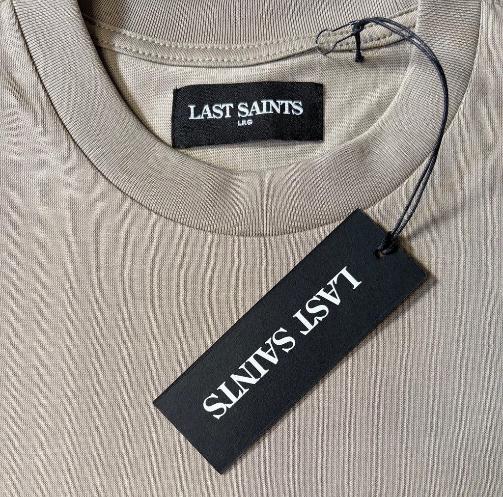 Close-up of a beige crewneck t-shirt with a black neck label reading 'LAST SAINTS LAW' and a black hangtag also printed with 'LAST SAINTS' in white, on a clean white background.