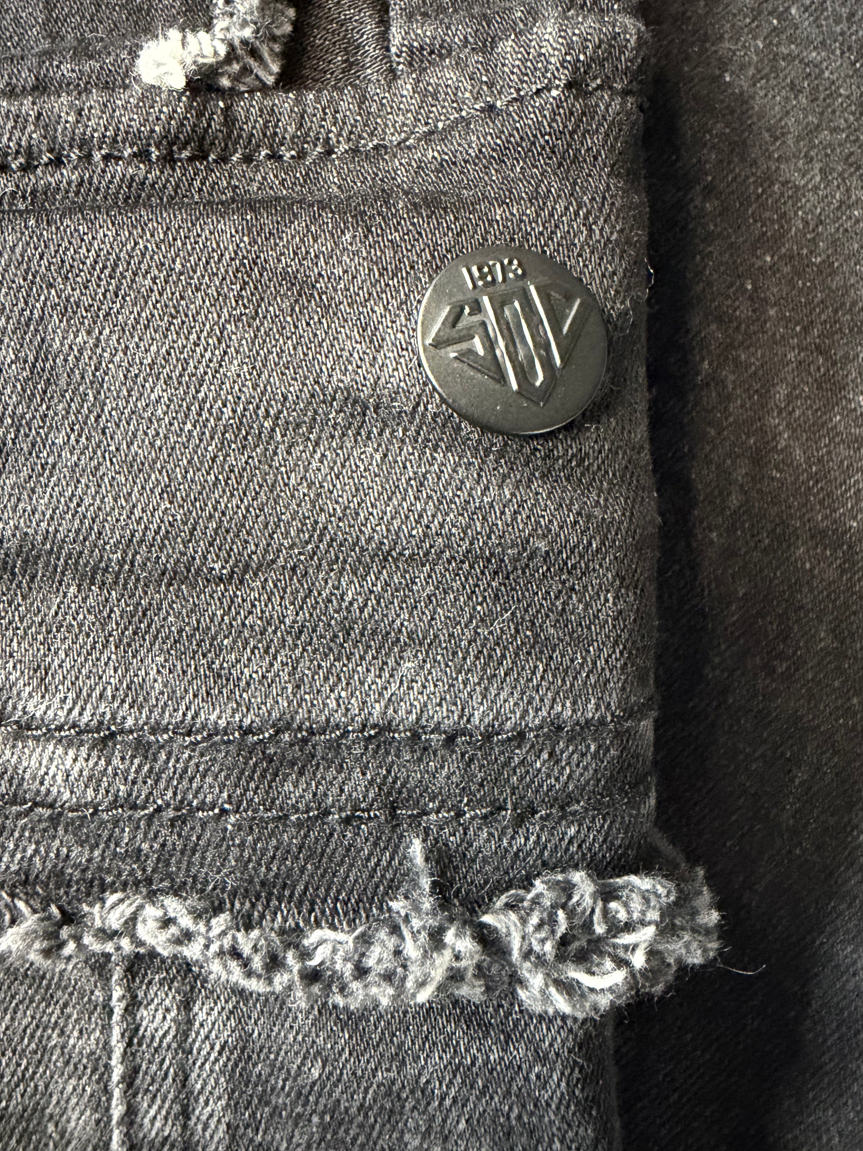 Detailed macro view of branded hardware on Stacked modern streetwear cargo jeans. Features a custom matte black metal shank button engraved with geometric logo text and a raw, distressed denim edge.