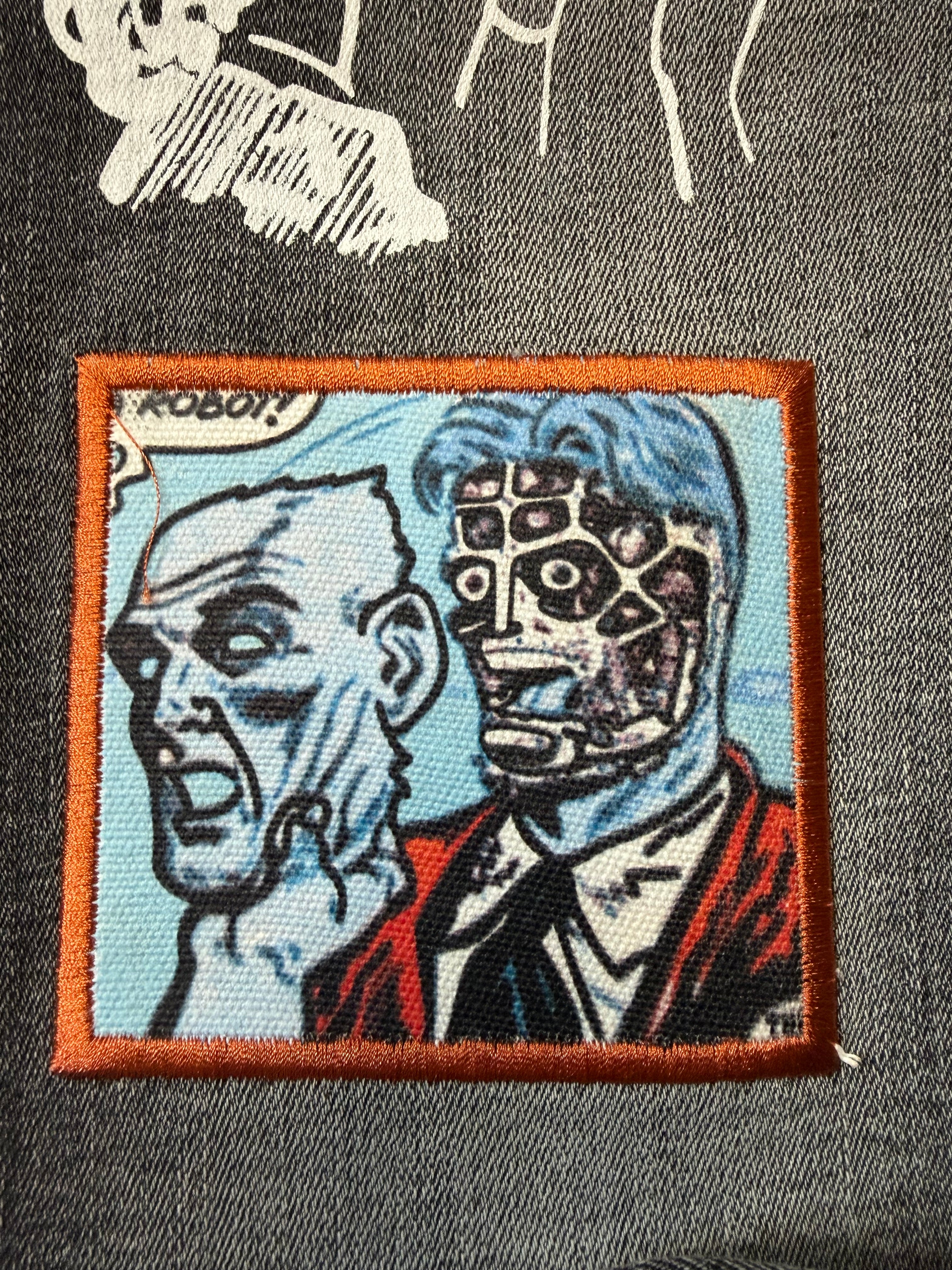 Macro detail view of an embroidered comic book illustration patch on Sugarhill streetwear jeans. Features retro pop art of a robot character with a speech bubble and dense orange border stitching.