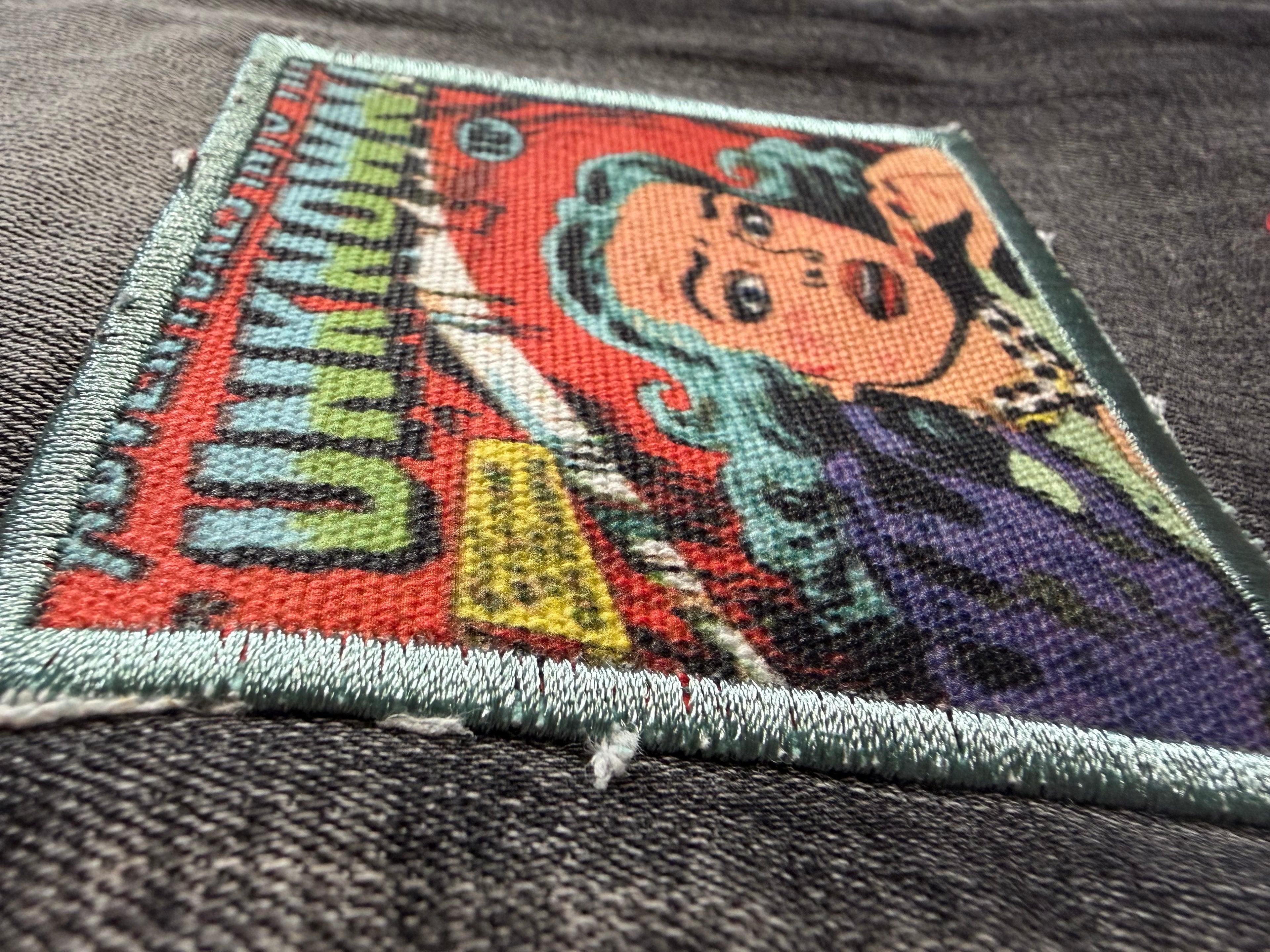 Macro detail shot of an embroidered comic book graphic patch on Sugarhill streetwear jeans. Features retro pop art of a woman with teal hair and vertical "UNKNOWN" lettering.