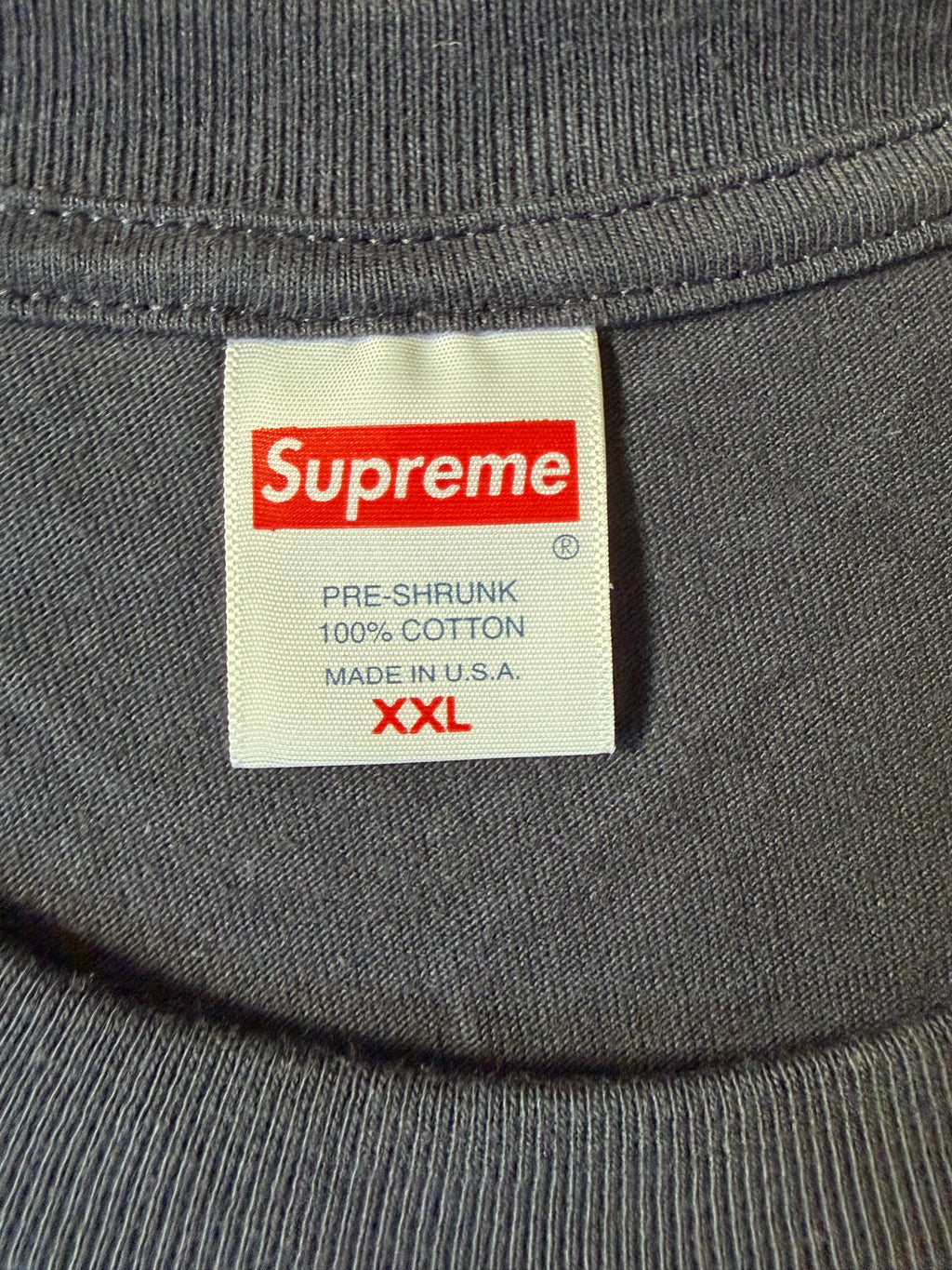 Close-up of a gray ribbed garment label with the red Supreme logo and text reading pre-shrink 100% cotton made in U.S.