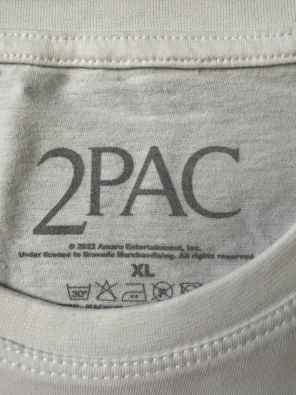 Close-up of the screen-printed inside neck collar of the official white Tupac unisex t-shirt, showing the dark gray 2Pac branding logo, size XL, and care instructions.